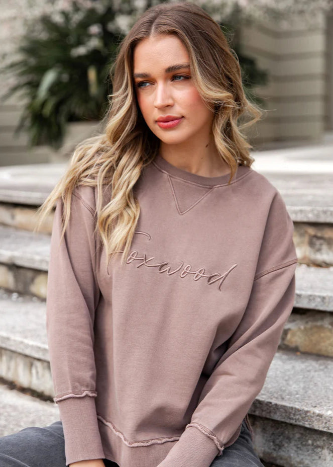 Woman wearing a mauve sweatshirt with 'Foxwood' text, sitting on stone steps.