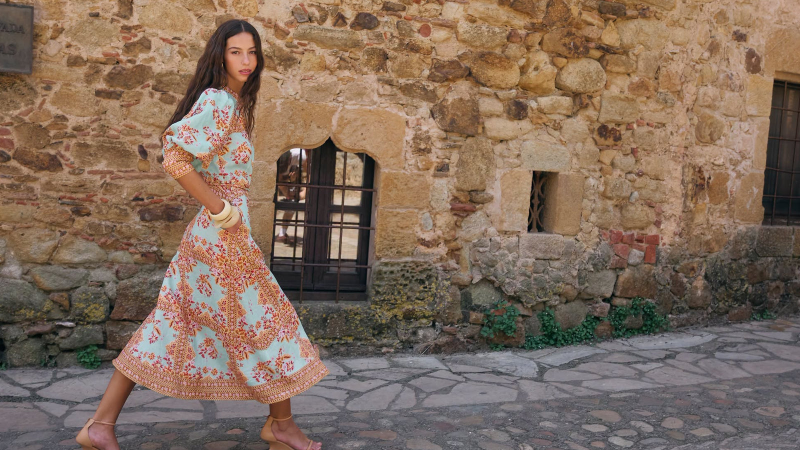 A woman wearing a light blue and orange floral midi dress walks past a rustic stone building with stone pavement.